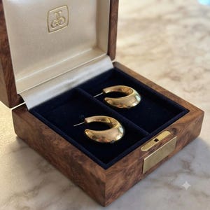 May include: A pair of polished gold hoop earrings with a thick, curved design resting inside a wooden jewelry box. The box features a dark blue velvet interior lining, a hinged lid with a gold logo emblem, and a brass latch on the front edge.