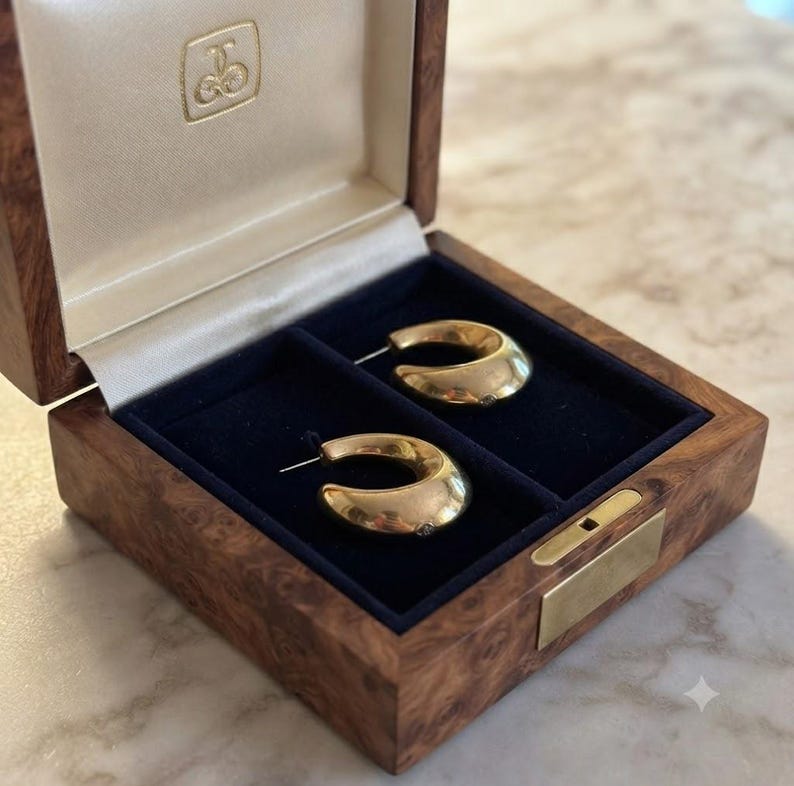 May include: A pair of polished gold hoop earrings with a thick, curved design resting inside a wooden jewelry box. The box features a dark blue velvet interior lining, a hinged lid with a gold logo emblem, and a brass latch on the front edge.