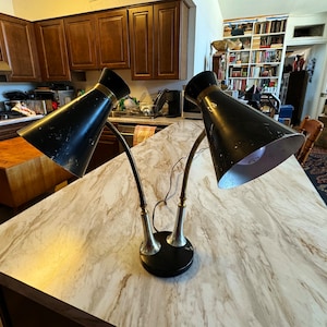 以下が含まれることがあります： 調節可能なアームと金のアクセントが付いた、黒いヴィンテージデスクランプ2つ。ランプは白い大理石のカウンタートップの上に置かれています。