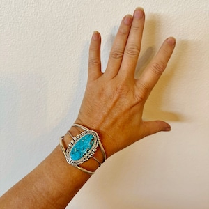 May include: A silver cuff bracelet with a turquoise stone set in the centre. The stone is oval shaped and has a dark blue and green colour.