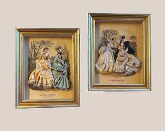 May include: Two framed vintage prints of women in dresses, set in gold-colored frames. The prints feature illustrations of women in elaborate gowns, with the text "LA MODE ILLUSTREE" at the bottom. The artwork is set against a neutral background.