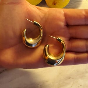 May include: A pair of gold-toned hoop earrings with a thick, chunky, crescent-shaped design. The earrings feature a smooth, reflective metallic surface and include a standard post-back closure mechanism for securing the jewelry to the earlobe.