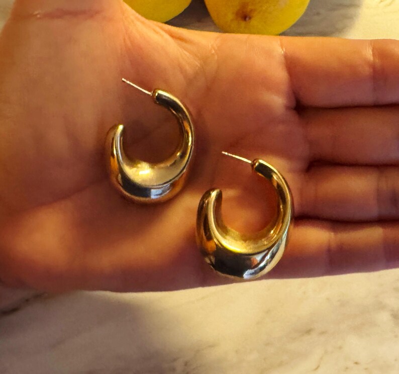 May include: A pair of gold-toned hoop earrings with a thick, chunky, crescent-shaped design. The earrings feature a smooth, reflective metallic surface and include a standard post-back closure mechanism for securing the jewelry to the earlobe.