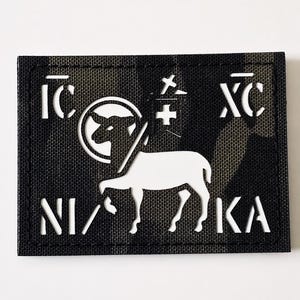 Può includere: Una toppa rettangolare con un motivo mimetico e simboli e testo bianchi. La toppa presenta un agnello, una croce e lettere greche. Il testo include "IC XC" e "NI KA".