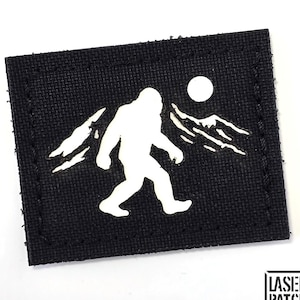 Könnte beinhalten: Schwarzer quadratischer Aufnäher mit einer weißen Silhouette von Bigfoot, der vor Bergen und einem Mond geht. Der Aufnäher hat einen genähten Rand und den Text "LASER PATCH" in einem Logo unten.
