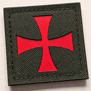 May include: A square-shaped patch featuring a dark olive green textured fabric border with visible stitching. Centered within the square is a bold, bright red cross cutout that reveals a smooth red base layer beneath the dark green top material.