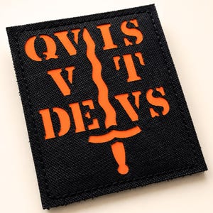 Könnte beinhalten: Schwarzer rechteckiger Aufnäher mit orangefarbenem Text und einem Schwertdesign. Der Text lautet "QVIS VT DEVS". Der Aufnäher hat einen genähten Rand und ist wahrscheinlich aus Stoff.