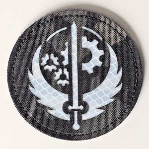 Puede incluir: Parche redondo con un patrón de camuflaje negro y gris. El parche presenta un emblema blanco reflectante con una espada, alas y formas de engranajes. El emblema está centrado en el parche.