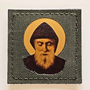 Könnte beinhalten: Quadratischer Aufnäher mit einem religiösen Bild, das einen goldenen Heiligenschein und eine bärtige Figur mit dunklem Hut zeigt. Das Bild befindet sich auf einem grünen Stoffhintergrund mit genähten Kanten.