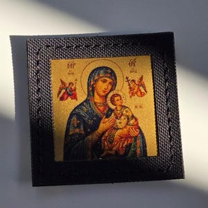 Könnte beinhalten: Ein quadratischer Aufnäher mit einer goldfarbenen religiösen Ikone der Jungfrau Maria, die das Jesuskind hält, umgeben von zwei kleinen Engeln. Das zentrale Bild ist von einem schwarzen, strukturierten Stoffrand mit sichtbaren Nähten umgeben.