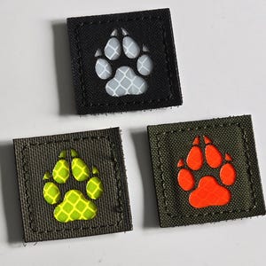 以下が含まれることがあります： 足跡のデザインが施された3つの正方形のパッチ。1つはグレーと白の足跡が付いた黒色、もう1つはネオンイエローの足跡が付いたオリーブグリーン、最後はオレンジ色の足跡が付いたオリーブグリーンです。すべてステッチされたボーダーがあります。