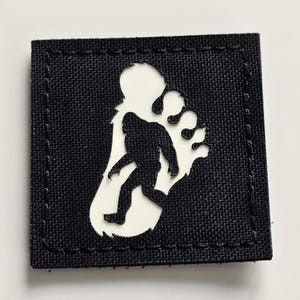 Può includere: Una toppa quadrata nera con un'impronta di Bigfoot bianca e una silhouette nera di Bigfoot. La toppa ha una superficie strutturata, probabilmente in tessuto. Il design è una rappresentazione della creatura mitica.