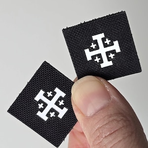 Könnte beinhalten: Zwei schwarze quadratische Aufnäher mit einem weißen Jerusalemer Kreuz-Design. Die Aufnäher werden vor einem weißen Hintergrund gehalten. Das Jerusalemer Kreuz ist ein heraldisches Symbol.