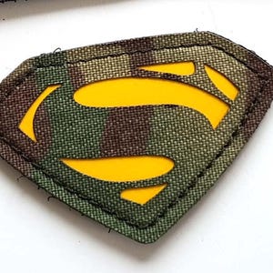 May include: Two embroidered patches. One black rectangular patch features three coloured lion silhouettes and the text "LASER PATCH" in white. The other patch is a camouflage diamond shape with a yellow "S" symbol.