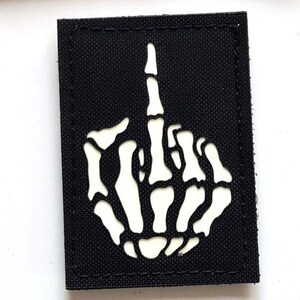 Könnte beinhalten: Zwei schwarze Aufnäher. Der obere Aufnäher hat ein Design von drei Löwen in Schwarz, Rot und Gelb, mit dem Text "LASER PATCH" in Weiß. Der untere Aufnäher zeigt eine weiße Skeletthand, die den Mittelfinger zeigt.