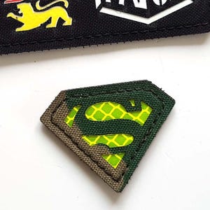 May include: Two embroidered patches. One is black with the text "LASER PATCH" in white and three coloured lion figures. The other is a green and brown Superman logo.
