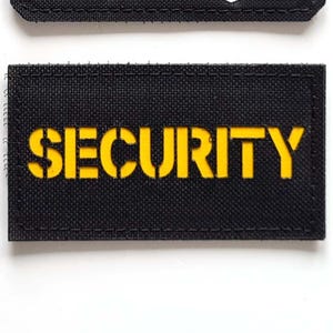 Può includere: Due toppe in tessuto nero. La toppa superiore presenta tre sagome di leoni in nero, rosso e giallo, con la scritta "LASER PATCH" in bianco. La toppa inferiore ha la parola "SECURITY" in giallo.