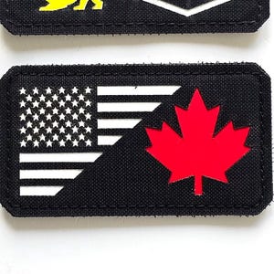 Peut inclure: Deux écussons rectangulaires noirs. L'un présente trois lions colorés et le texte "LASER PATCH" en blanc. L'autre affiche le drapeau américain et la feuille d'érable canadienne.