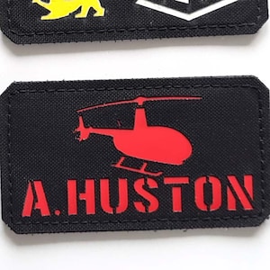Puede incluir: Dos parches rectangulares negros. El parche superior tiene tres figuras de leones en negro, rojo y amarillo, con el texto "LASER PATCH" en blanco. El parche inferior presenta un helicóptero rojo y el texto "A. HUSTON" en rojo.