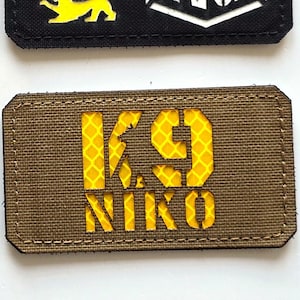 Op de afbeelding: Twee rechthoekige patches. De bovenste patch is zwart met drie leeuwsilhouetten in zwart, rood en geel, en de tekst "LASER PATCH" in wit. De onderste patch is bruin met de tekst "K9 NIKO" in geel.