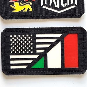 Może przedstawiać: Dwie czarne prostokątne naszywki. Górna naszywka ma trzy kolorowe lwy i napis "LASER PATCH". Dolna naszywka przedstawia flagę amerykańską i flagę włoską.