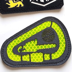 Puede incluir: Dos parches bordados. El parche superior es rectangular, negro, con tres figuras de león y el texto "LASER PATCH" en blanco. El parche inferior tiene forma de mosquetón, gris, con un patrón de rejilla verde neón.