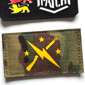 Może przedstawiać: Dwie haftowane naszywki. Górna naszywka jest czarna z trzema kolorowymi figurami lwa i napisem "LASER PATCH" w kolorze białym. Dolna naszywka jest moro z żółtą błyskawicą i gwiazdami.