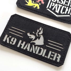 May include: Two black rectangular patches. One patch features three lion figures in grey, red, and yellow, and the text "LASER PATCH" in white. The other patch has a dog silhouette and the text "K9 HANDLER" in white.