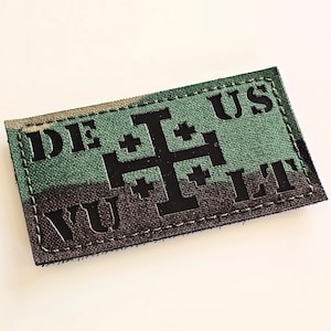 May include: A rectangular patch featuring a two-tone green and dark brown camouflage pattern. The centre displays a black Jerusalem cross symbol, flanked by the black text DEUS VULT, with DE US positioned above the cross and VU LT below it.