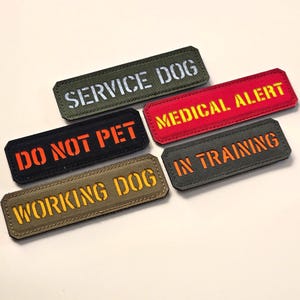 Może przedstawiać: Pięć prostokątnych naszywek z tkaniny z wyhaftowanym tekstem. Oliwkowozielona naszywka ma napis SERVICE DOG, czerwona MEDICAL ALERT, czarna DO NOT PET, beżowa WORKING DOG, a szara IN TRAINING.