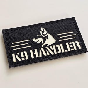 Può includere: Toppa rettangolare nera con testo bianco e sagoma di cane. Il testo recita "K9 HANDLER" sotto una grafica a testa di cane e due linee orizzontali. La toppa è probabilmente realizzata in tessuto resistente.