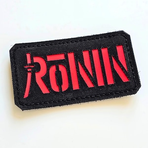 Puede incluir: Parche rectangular negro con la palabra "RONIN" en rojo, con letras japonesas estilizadas. El parche tiene un borde negro con bordes cosidos. El fondo es blanco.