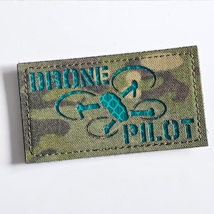 May include: A rectangular patch with a camouflage pattern in shades of green and brown. The patch features the words "DRONE PILOT" in teal, along with a teal drone graphic. The patch has a black border and is likely made of fabric.