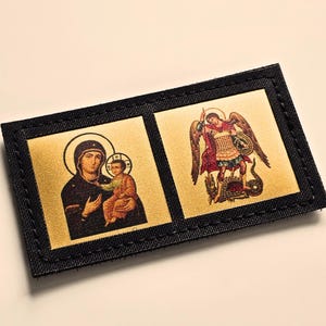 Puede incluir: Un parche rectangular con borde de tela negra que presenta dos paneles dorados. El panel izquierdo muestra un icono de una figura sosteniendo a un niño, mientras que el derecho representa una figura alada con armadura sobre una criatura serpentina.