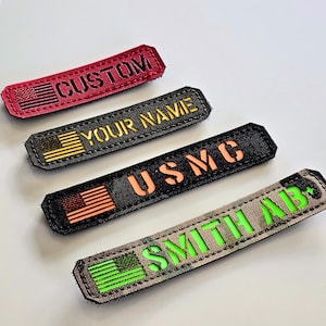 May include: Four rectangular patches with embroidered text and American flag designs. The patches are in red, black, and camouflage patterns. Text includes "CUSTOM", "YOUR NAME", "USMC", and "SMITH AB".