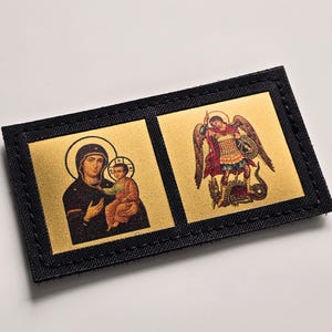 May include: A rectangular patch with two gold squares displaying religious icons. One square shows a woman and child, the other an angel. The patch has a black border with visible stitching around the edges.
