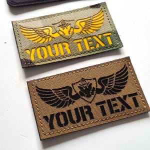 Könnte beinhalten: Zwei rechteckige Aufnäher mit gestickten Designs. Einer ist Camouflage mit gelbem Text und einem geflügelten Emblem. Der andere ist beige mit schwarzem Text und einem ähnlichen Emblem. Beide lauten "YOUR TEXT".