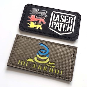 May include: Two rectangular patches. One black patch features three lion emblems in silver, red, and yellow, and the text "LASER PATCH" in white. The second patch is olive green with a blue and yellow snake design and the text "IDI-NAKHUI".