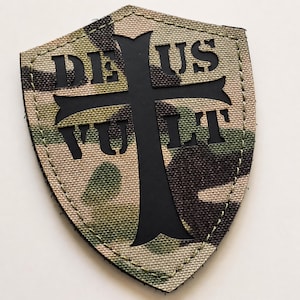 Könnte beinhalten: Ein schildförmiger Aufnäher mit einem Tarnmuster in Beige-, Grün- und Brauntönen. Ein großes schwarzes Kreuz ist mittig auf dem Aufnäher platziert, wobei der schwarze Schriftzug DE US VULT in das Design integriert und um die Kreuzarme angeordnet ist.