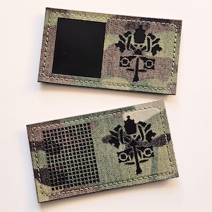 Könnte beinhalten: Zwei rechteckige Aufnäher mit Tarnmuster in Grün-, Braun- und Beigetönen. Jeder Aufnäher zeigt ein schwarzes Emblem mit gekreuzten Schlüsseln und einer Tiara. Einer hat ein schwarzes Quadrat, der andere ein schwarzes Gittermuster.