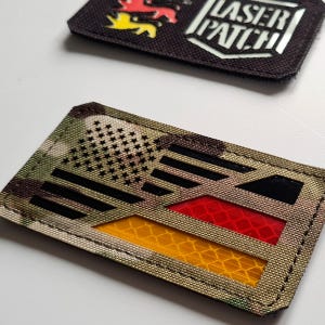 Può includere: Due toppe tattiche. Una presenta un design a bandiera americana mimetica con accenti neri, rossi e gialli. L'altra è nera con uno stemma e le parole "LASER PATCH" in bianco.