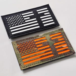 May include: Two rectangular patches featuring the American flag design. One patch is black with white stripes and stars. The other patch has a camouflage background with orange stripes and stars. Both patches have a stitched border.