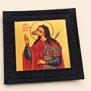 Pode incluir: Um emblema quadrado com um ícone de São Cristóvão com cabeça de cão, vestindo roupas vermelhas e azuis e segurando uma lança. A imagem está sobre um fundo dourado e emoldurada por uma borda de tecido preto texturizado com costuras.
