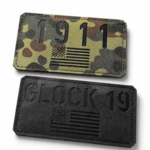 Könnte beinhalten: Zwei rechteckige Aufnäher mit Klettverschluss auf der Rückseite. Der obere Aufnäher zeigt ein grünes, braunes und schwarzes Tarnmuster mit dem schwarzen Text 1911 und einer kleinen amerikanischen Flagge. Der untere Aufnäher ist schwarz mit dem Text GLOCK 19.