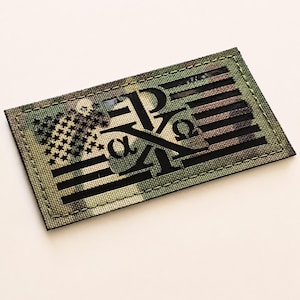 May include: A rectangular morale patch featuring a camouflage pattern in shades of green, brown, and black. The design incorporates a stylized American flag with stars and stripes, overlaid with the Greek letters Chi, Rho, Alpha, and Omega in black.