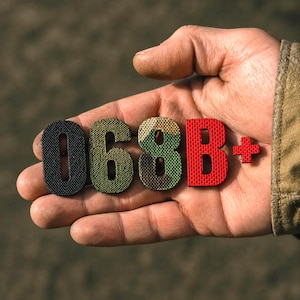 Könnte beinhalten: Eine Hand, die Stoffbuchstaben und ein Pluszeichen hält. Die Buchstaben ergeben "068B+". Die "0" ist schwarz, die "6" und "8" sind Tarnmuster, das "B" ist rot und das Pluszeichen ist rot. Verschwommener Hintergrund.