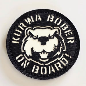 May include: A round, black patch with white text and a white beaver illustration. The text reads "KURWA BOBBER ON BOARD!" in a bold, sans-serif font. The beaver has an angry expression.