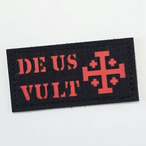 May include: A black rectangular patch featuring the words "DEUS VULT" in red, alongside a red Jerusalem cross. The patch is likely made of fabric and has stitched edges. The text is in a bold, sans-serif font.