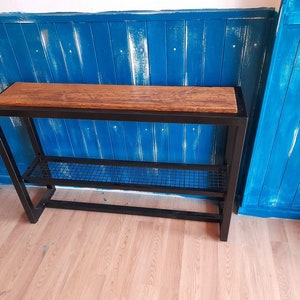 May include: A black metal console table with a wooden top and a wire shelf. The table is against a blue wall.