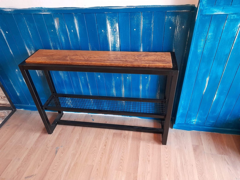 May include: A black metal console table with a wooden top and a wire shelf. The table is against a blue wall.